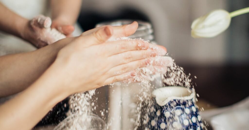 Hands of parent and child baking together, scattering flour on a wooden board with eggs and teapot nearby.